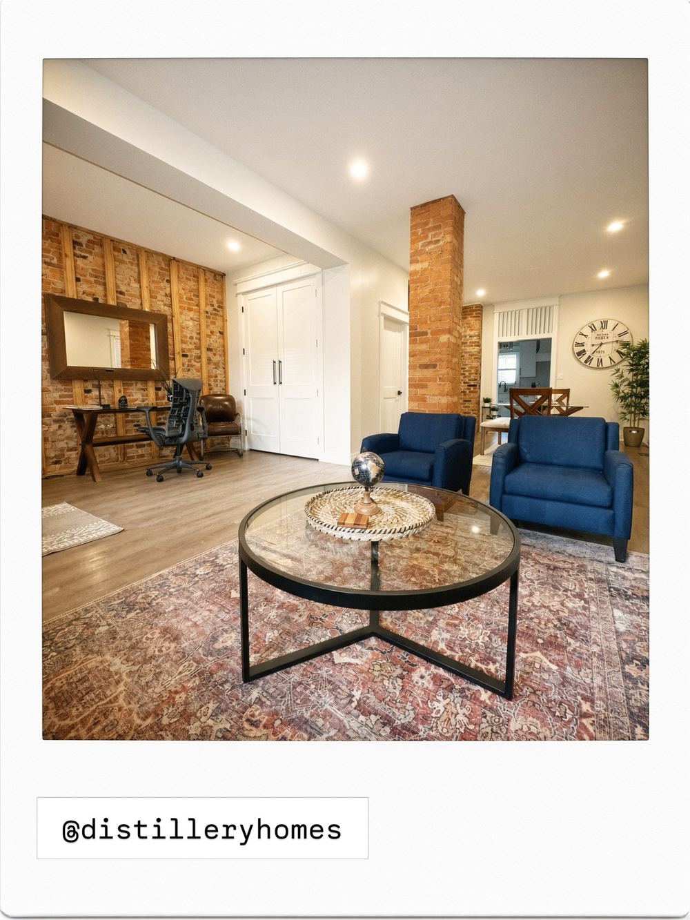 Open-concept living room at 763 Monmouth Road, Walkerville, with exposed brick walls and chimney column, navy armchairs, round glass coffee table and a home office nook